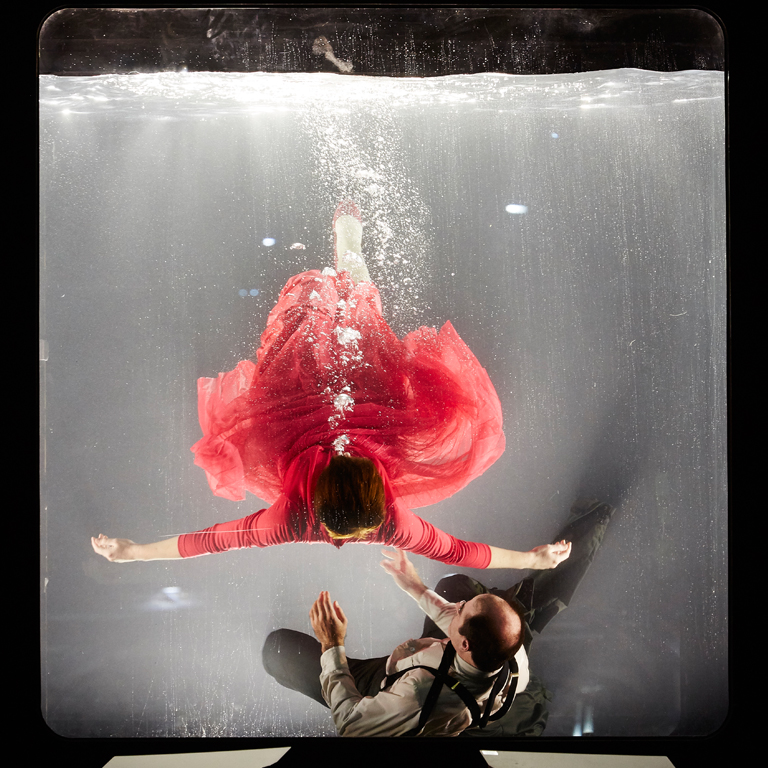 Woman in a red dress and man in a suit in a clear cube tank filled with water