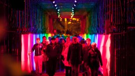 People walking through a square tunnel lit up in rainbow colours