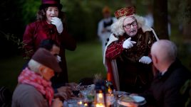 People sat a dining table are looking at a man pointing at one of the people seated. He is dressed as a King with a crown, a white painted face and pink circles on his cheeks