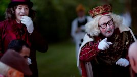 People sat a dining table are looking at a man pointing at one of the people seated. He is dressed as a King with a crown, a white painted face and pink circles on his cheeks