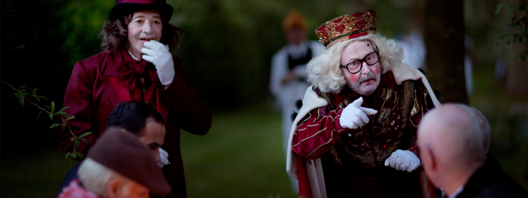 People sat a dining table are looking at a man pointing at one of the people seated. He is dressed as a King with a crown, a white painted face and pink circles on his cheeks