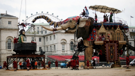 Side view of giant elephant puppet facing giant girl puppet sitting in a black bowl. Part of the London Eye is visible from in the background