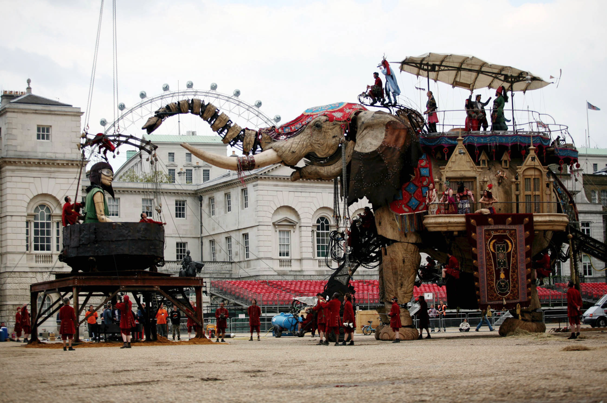 Side view of giant elephant puppet facing giant girl puppet sitting in a black bowl. Part of the London Eye is visible from in the background