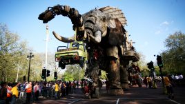 Giant puppet The Sultan's Elephant picking up a yellow pick up truck with its trunk with a crowd around it.