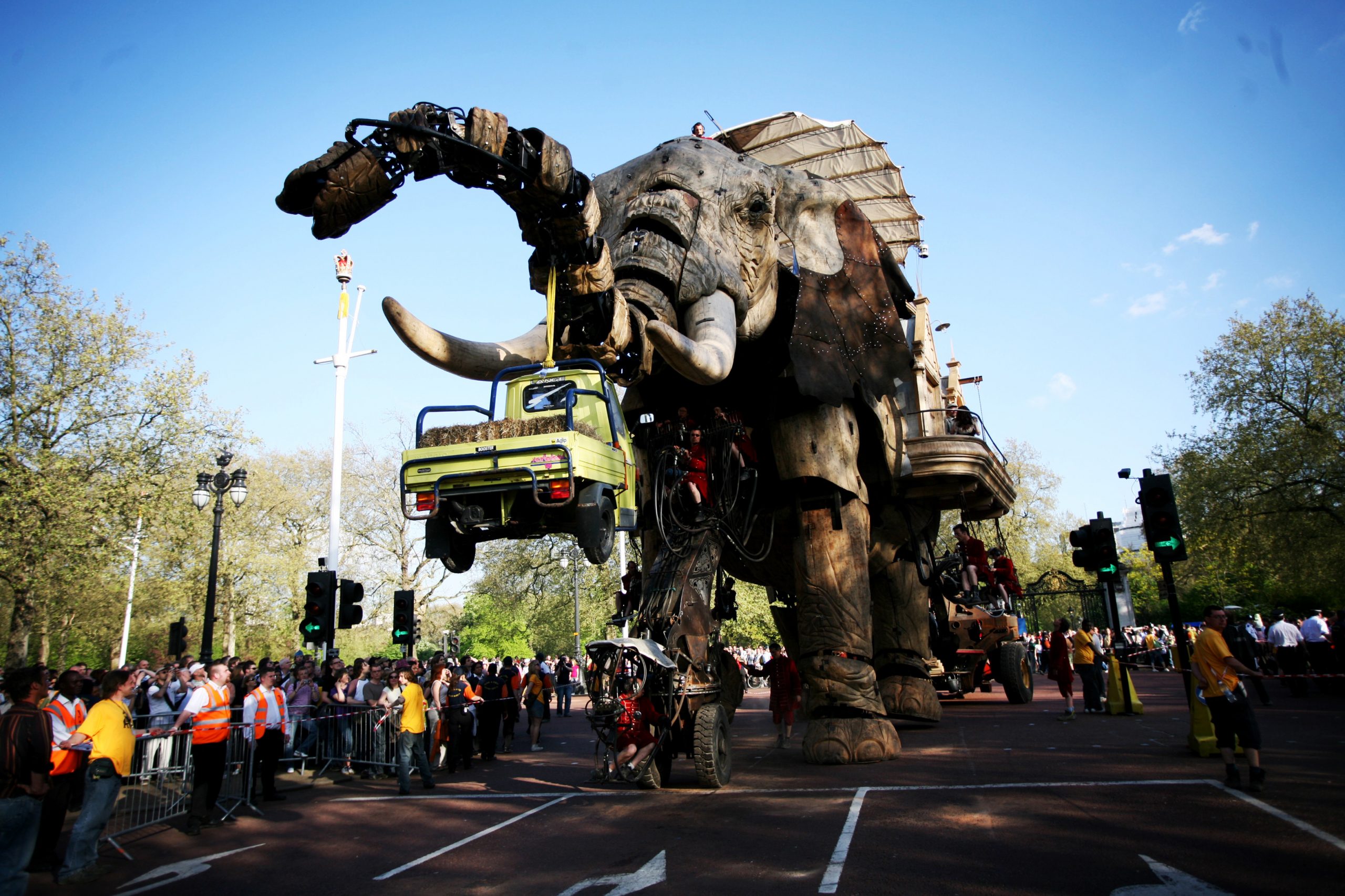 Giant puppet The Sultan's Elephant picking up a yellow pick up truck with its trunk with a crowd around it.