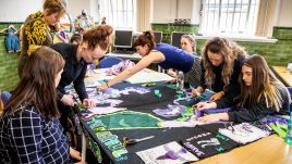 Seven women are leading across a table. They are talking and working with paper and materials. They look happy and engaged in the task.