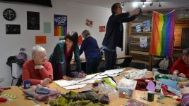 Three women are sitting at a table working with materials and paper. A man in the background is hanging up a rainbow coloured flag.