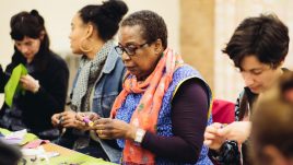 A woman wearing a blue top and an orange scarf holding a cotton reel with purple thread. She looks engaged in the activity.