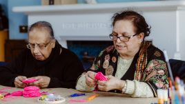 A close-up of two women sitting at a table sewing small pieces of pink fabric. They look happy and engaged.