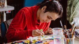 A woman wearing a red jumper is sitting at a table making a print on paper with yellow ink.