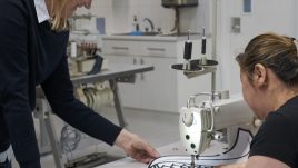 Artist Lucy is wearing a shirt and a jumper. She has blonde hair. She is leading towards a woman sitting at a sewing machine and talking to her. They both look engaged in the conversation.