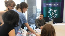 Rudy is wearing a shirt and glasses and they are sitting at a table talking about ideas for the banner design with women from the group.