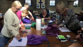 Women in a church standing around a table and working together to sew pieces of brightly coloured material. They look happy and engaged in the task.