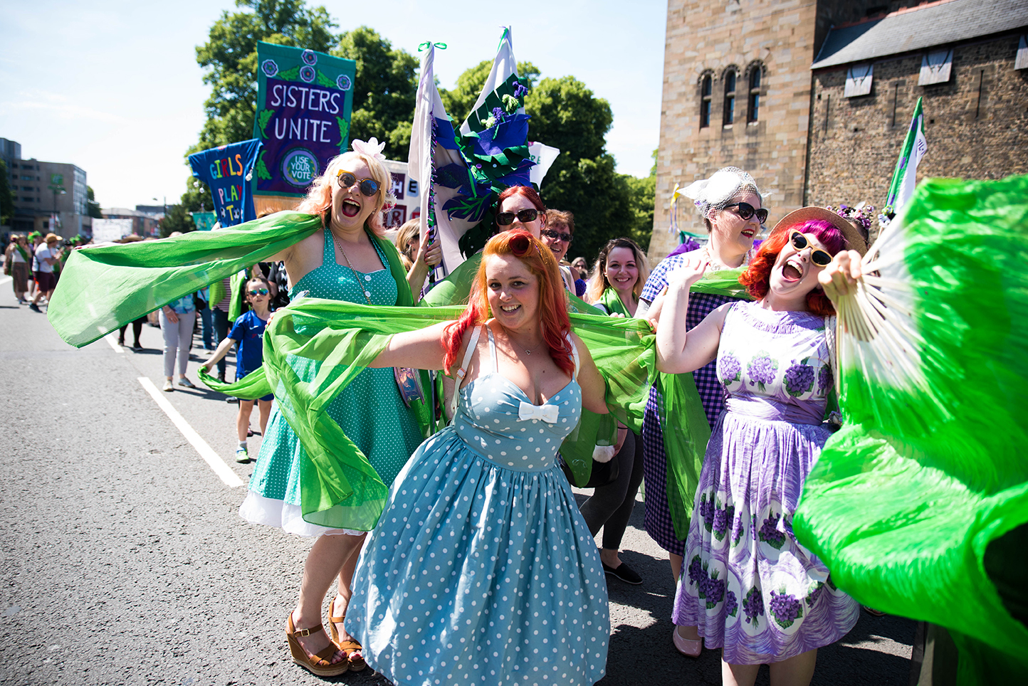 Group of women with green sashes and a green fan smiling at the camera in the midst of the Procession in Cardiff
