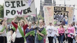 A crows at PROCESSIONS in Edinburgh. Women hold a banner above their heads. Text reads: Girl power