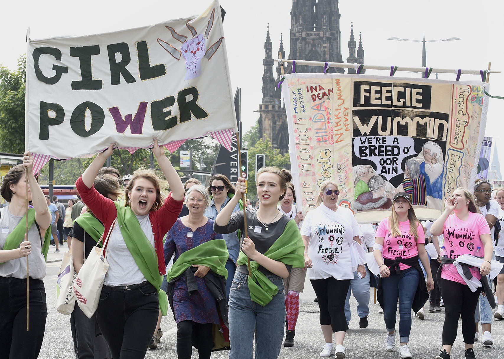 A crows at PROCESSIONS in Edinburgh. Women hold a banner above their heads. Text reads: Girl power