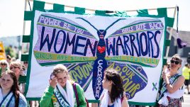 Women holding up Green and purple banner with a purple butterfly in the centre. On the banner is written WOMEN WARRIORS and other inspiration phrases
