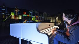 Man playing piano in foreground and light installation illuminating a building.