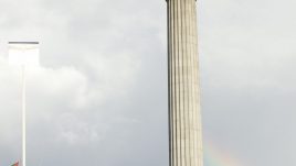 Man standing stop the plinth in a suit with rainbow in the background.