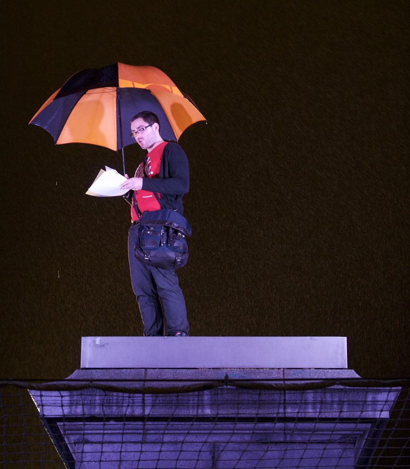 Man holding a black and red umbrella reading from sheets of paper while standing on the fourth plinth for One & Other