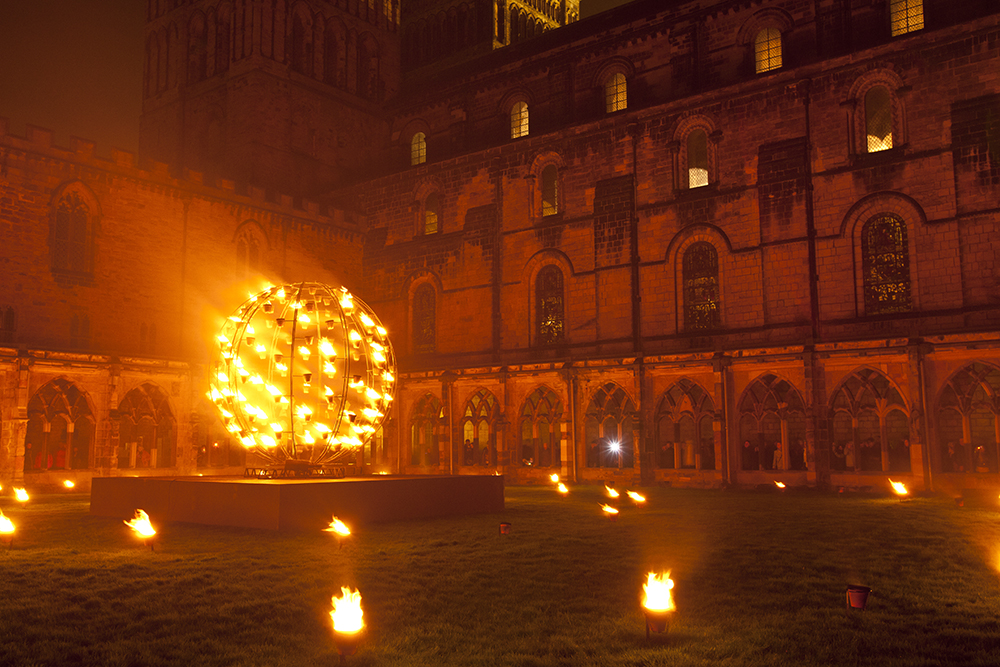 A metal ball with flames surrounded by smaller flames on the ground outside Durham Cathedral