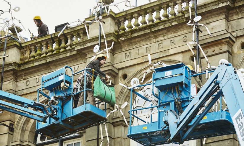 Men in cherry pickers installing the light work Key Frames on Durham Miner's Hall