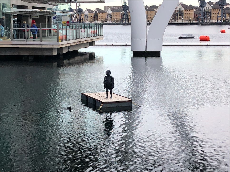 In the middle of the river, on a floating wooden square is a sculpture of the silhouette of a young boy with the face of a bird