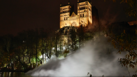 View of the cathedral from the River Wear at dusk. The Cathedral is lit up from belong. Above the river below the catherdral is a big cloud of white smoke.