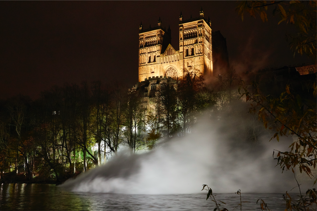 View of the cathedral from the River Wear at dusk. The Cathedral is lit up from belong. Above the river below the catherdral is a big cloud of white smoke.