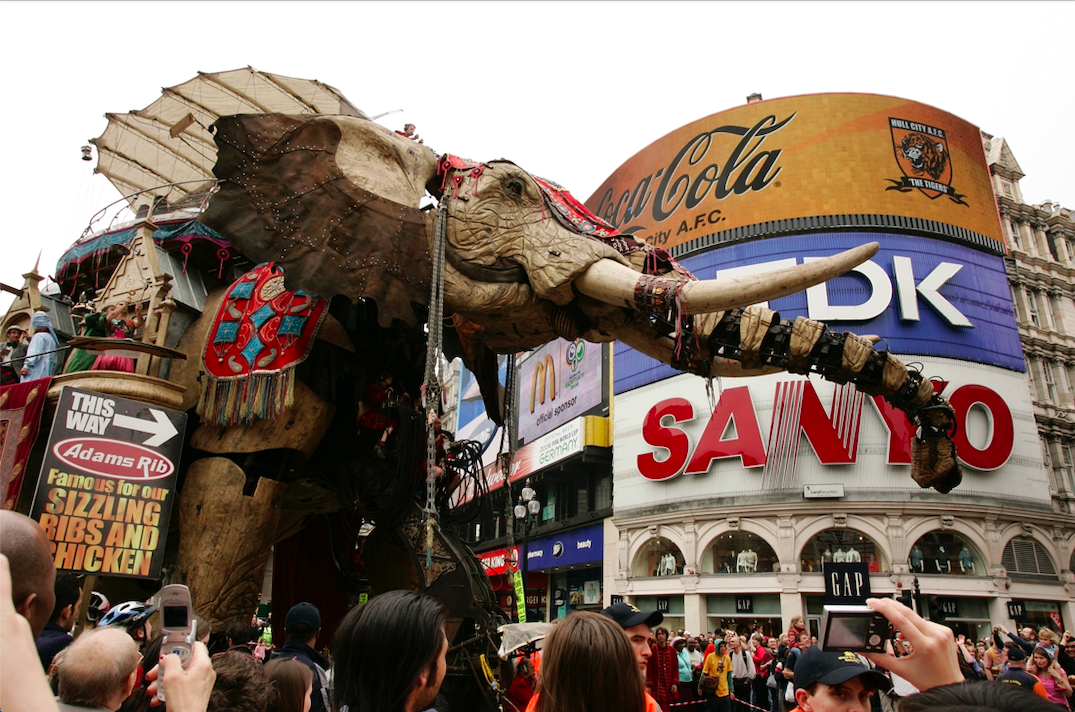 A below view of a giant puppet elephant in front of advertising signage with a large crowd around it.