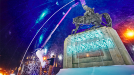 Man inside the snowglobe, empty a large bag with the fake snow onto the ground. Behind him is the statue of a man riding a horse on a plinth with the illuminated words I LOVE DURHAM on it.