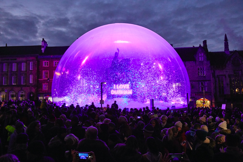 A large crowd gathered around a snow globe, in the snow globe is glitter and at the centre is a statue of a man riding a horse on a plinth with the illuminated words I LOVE DURHAM on it.