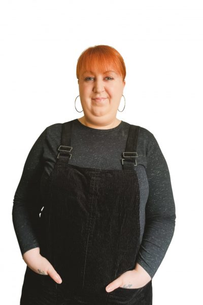 Headshot of Steph smiling, she's wearing hoop earrings, a grey top and a black dungaree dress