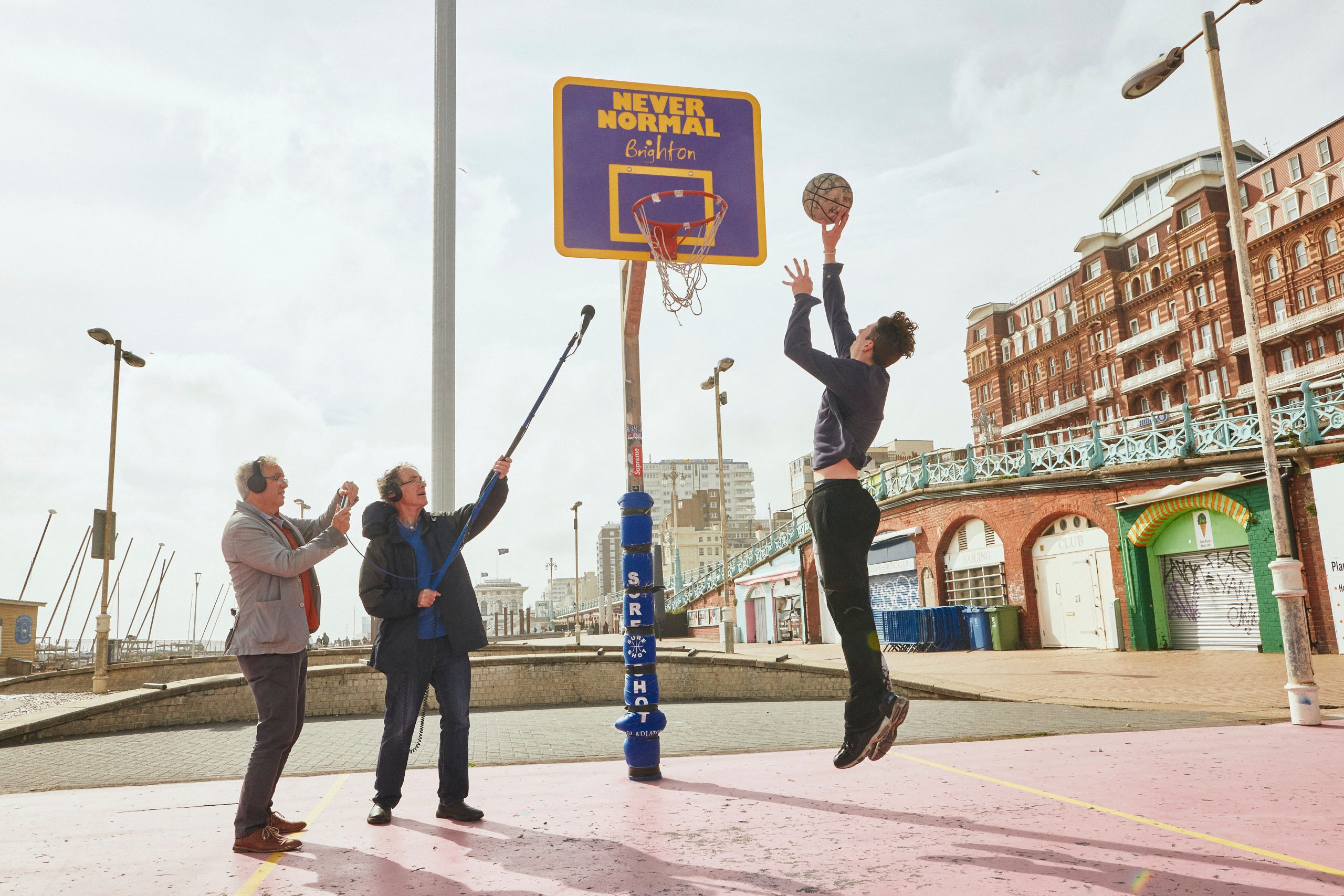 Man jumping in the air about to dunk a basketball into the net, on the right are two men one of which is holding up a microphone recording the sounds