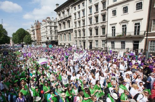 A still of the crowd of women walking in London during PROCESSIONS, in a wave of green white and purple