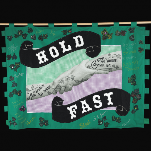 Slogan “HOLD FAST” on a banner, in the center is an image of two arms clasping at each other, both are tattooed, one with the words “The women before us