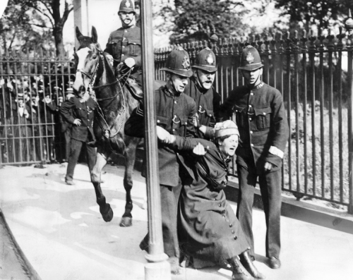 black-and-white image of a woman campaigning for the vote being restrained by three policemen, behind them is a fourth policeman on a horse