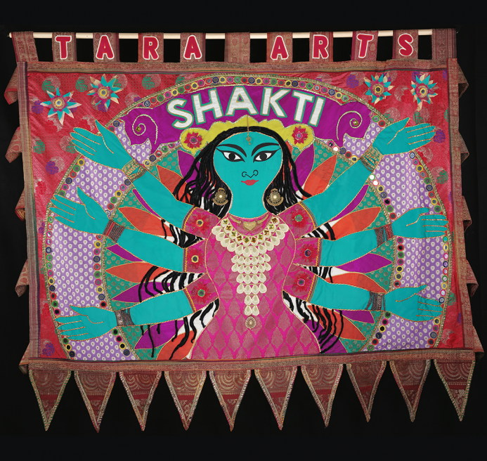 Blue human figure with 6 arms against pink and orange background made from recycled saris. SHAKTI is written above its head.