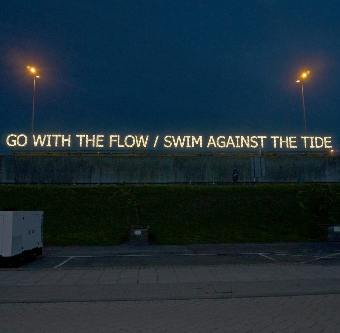 GO WITH THE FLOW / SWIM AGAINST THE TIDE neon sign above a carpark
