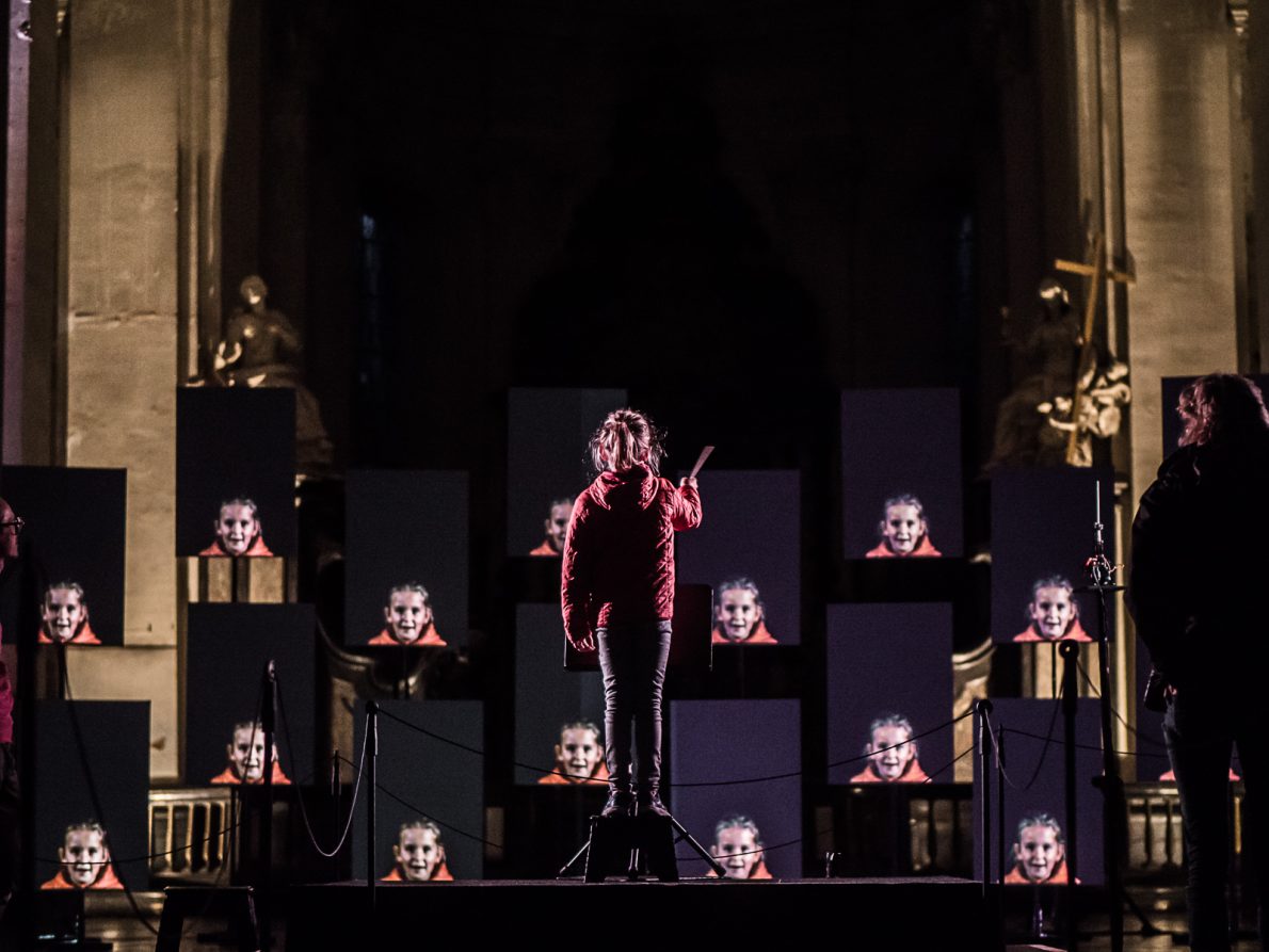 young girl stood up conducting and in front of her are multiple screens with an image of her face live