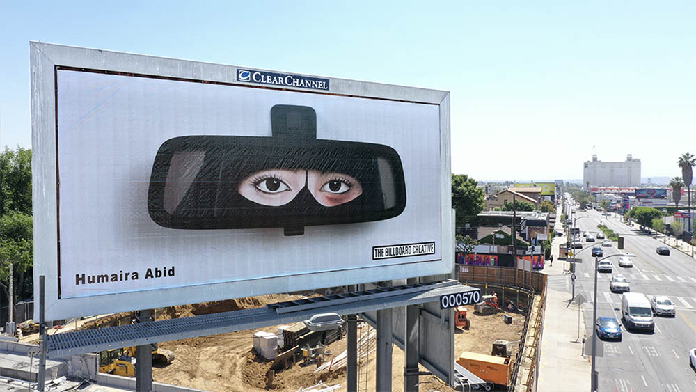 A billboard above a busy highway in LA. The artwork is of a car mirror with the reflection of woman wearing a burka.