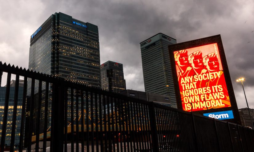 Red billboard in front of a cityscape featuring the words 'Any society that ignores its own flaws is immoral'