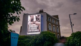 Photo of art by Martin Firrell on a digital screen. The image depicts a close up black and white photo of a commuter's crotch. The printed writing reads; 