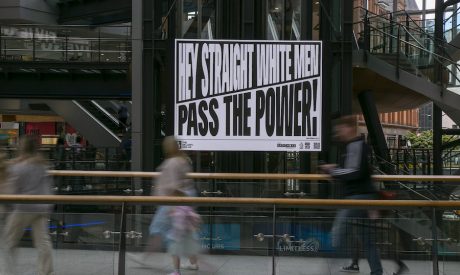 Text artwork on an outdoor digital screen. Text reads: 'Hey Straight White Men, Pass the Power'