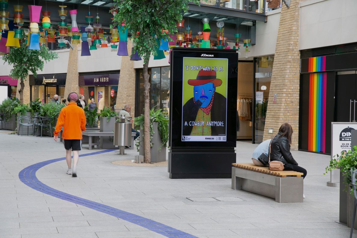 A digital ad screen displays the artwork 'I Don’t Want To Be A Cowboy Anymore' from Season 1 of The Gallery. A blue cowboy nervously holding his hand in front of his mouth. Text reads: 'I don't want to be a cowboy anymore.'