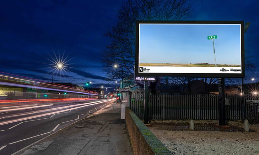 A billboard of CRY (2022) by Allyson Packer overlooking a busy road at night. A barren landscape. A freight train can barely be seen in the distance. A road sign in the foreground reads 'CR Y'.