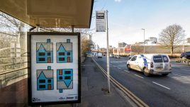 A bus shelter screen of the artwork 'A house upside down’ (2023) by Richard Woods in Manchester. Four geometric blue houses with grey roofs and orange doors. Three stand upright and one is upside down.