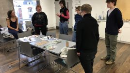A photo from a workshop in Belfast for Season 2 of The Gallery. A group of young people stand in a room with piles of art supplies on their work stations. They are creating artwork in repsonse to the theme 