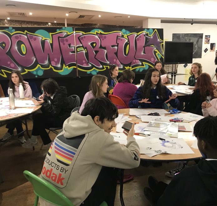 A photo from a schools workshop in Cardiff for Season 2 of The Gallery. A group of young people sit in a classroom with piles of art supplies on their work stations. They are creating artwork in repsonse to the theme 