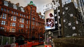 A billboard of the artwork 'Care' by Dola Posh on a busy London road. A black woman sits atop her bed with a toddler sat cross-legged in front of her. She is wearing a floral robe with a burgundy head wrap. The woman combs out her daughter's afro and puts it into twists.